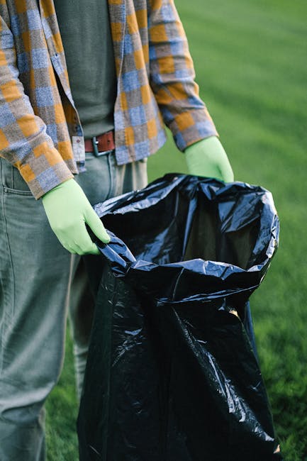 A worker in a red and yellow high-visibility jacket is operating a large red rubbish collection vehicle on the side of a road, with the rear doors open for loading waste. The worker is positioned near