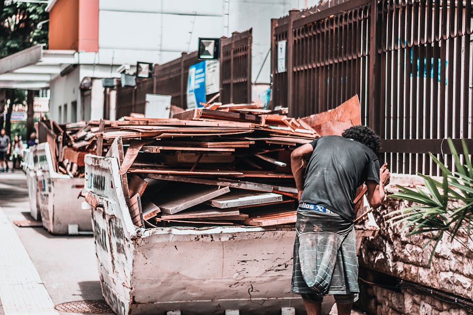 A white rubbish collection truck is parked on a city street in front of a multi-storey brick building with large, grid-pane windows, some of which are open. The truck is heavily loaded with various ty