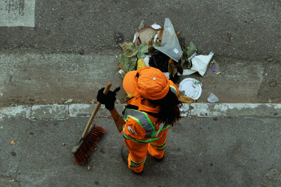 An aerial view of a bright green waste collection vehicle parked on a city street, with its rear hatch open, revealing collected black and transparent plastic rubbish bags filled with mixed waste. The