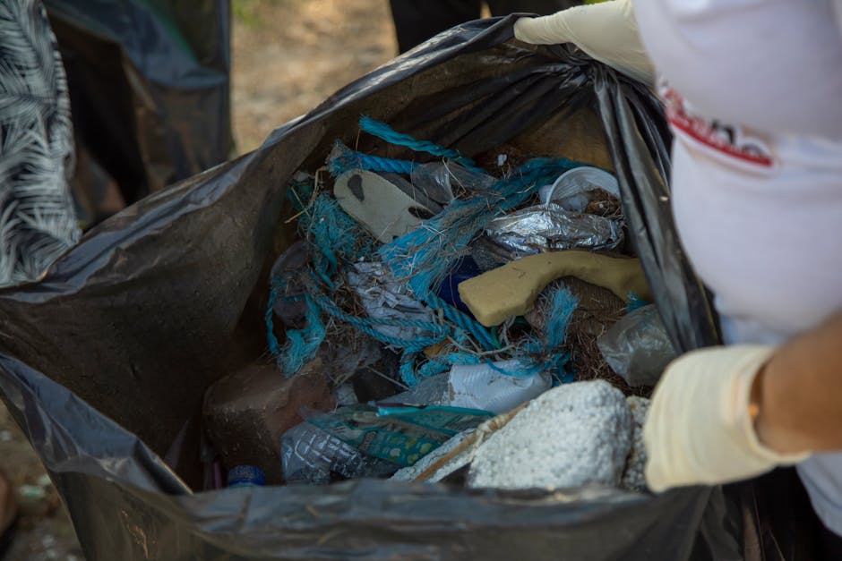 A close-up view of a black plastic rubbish bag filled with mixed waste, including torn blue plastic ropes, crumpled aluminum foil, a yellow rubber glove, a piece of white foam or insulation, and vario