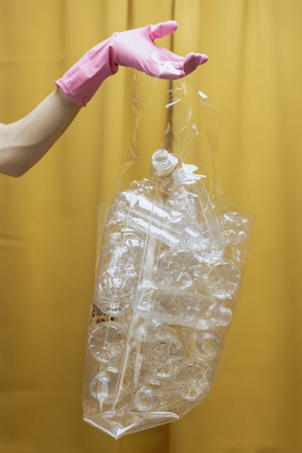 A close-up view of a black plastic rubbish bag filled with mixed waste, including torn blue plastic ropes, crumpled aluminum foil, a yellow rubber glove, a piece of white foam or insulation, and vario