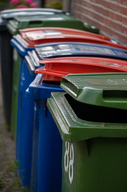 A row of large, rectangular wheelie bins with hinged lids, positioned on a paved sidewalk beside a brick wall. The bins are primarily blue, green, and red, with the green bin in the foreground featuri
