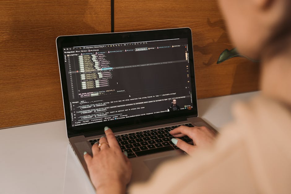 A person with short, light-colored nail polish and a ring on their left hand types on a MacBook laptop that displays lines of programming code in a dark-themed editor. The laptop is placed on a white 